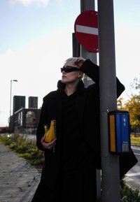 Svart oversized jacka, svart huvtröja och solglasögon. Håller en liten gul handväska med kedjestrap. Står nära ett trafikskylt och stolpe.