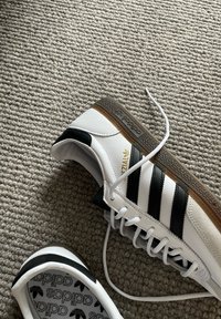 Baskets Adidas blanches avec des bandes noires et des semelles marron sur un tapis beige texturé, une chaussure inclinée montrant le logo de la semelle intérieure.