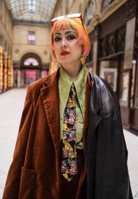 Blazer di velluto marrone sopra una camicia verde con motivo, con cravatta colorata con stampa di auto d'epoca. Capelli corti con sfumature rosa e arancioni.