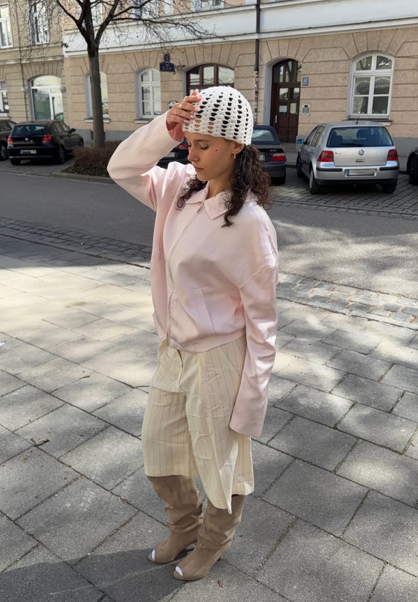 Giovane donna con giacca rosa chiaro, pantaloni crema a righe, stivali beige e cappello bianco lavorato a maglia, in piedi sul marciapiede della città, toccandosi la testa.