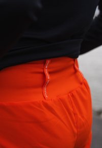 Φωτεινά πορτοκαλί αθλητικά σορτς με μαζεμένη μέση και ορατή λευκή ετικέτα στη ζώνη που αναγράφει "SOUND BODY". Λείος, ελαφρύς ύφασμα.