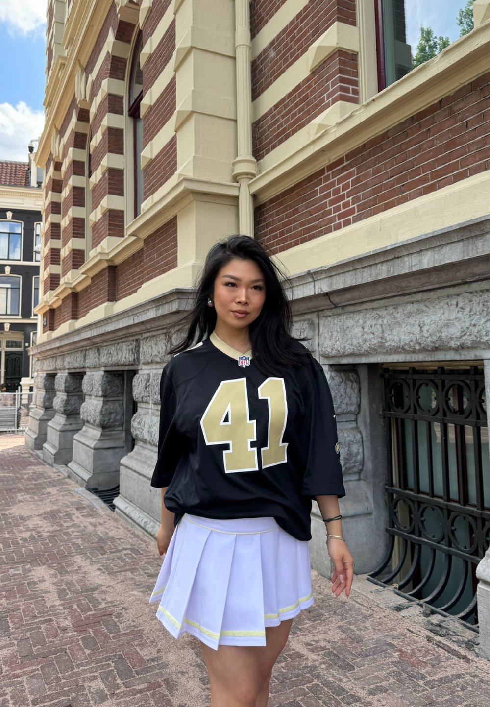 Maglia da calcio nera con numeri e logo dorati, abbinata a una gonna bianca plissettata con un bordo giallo accentato. Ambientazione urbana con sfondo in mattoni.