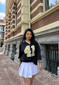 Maglia da calcio nera con numeri e logo dorati, abbinata a una gonna bianca plissettata con un bordo giallo accentato. Ambientazione urbana con sfondo in mattoni.