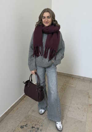 Mujer con suéter gris, jeans de pierna ancha, zapatillas blancas y gran bufanda burdeos oscuro sosteniendo un bolso a juego en el interior sobre suelo de baldosas.