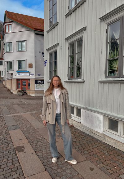 Trench beige claro sobre una camisa blanca, combinado con pantalones vaqueros de pierna ancha azul claro y zapatillas blancas. Calle de adoquines y edificios de madera al fondo.