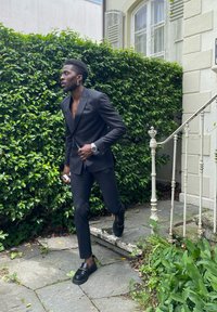 Zwart slim-fit pak met spitse revers, open kraag en zwarte loafers. Zilveren horloge en oorbellen. Natuurlijke groene bladeren en stenen trappen op de achtergrond.