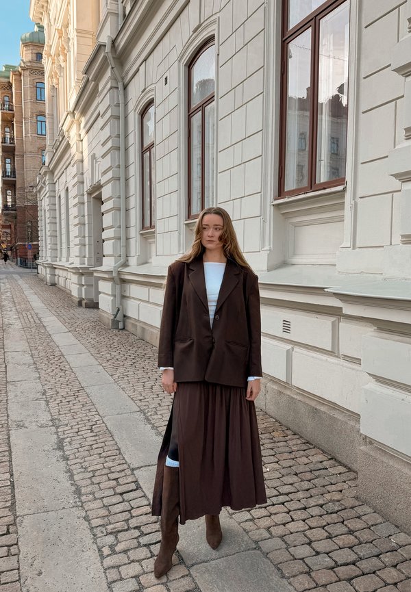 Žena u smeđem blejzeru, dugoj plisiranoj suknji i čizmama stoji na kaldrmisanoj ulici pored bijele kamene zgrade s velikim prozorima.
