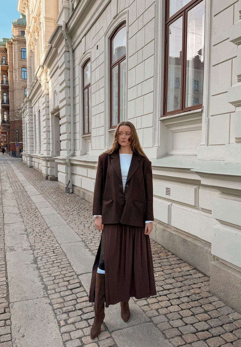 Femme portant un blazer marron, une longue jupe plissée et des bottes, debout dans une rue pavée à côté d'un bâtiment en pierre blanche avec de grandes fenêtres.