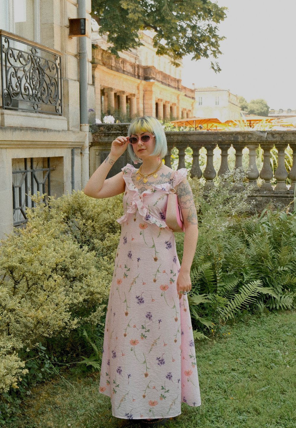 Rosa blomster maxi-kjole med rynket halsudskæring, prydet med livlige blomster mønstre. Modellen bærer solbriller og holder en pink håndtaske. Grønne blade i baggrunden.