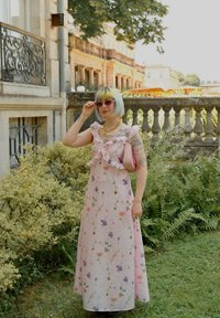 Rosa blomster maxi-kjole med rynket halsudskæring, prydet med livlige blomster mønstre. Modellen bærer solbriller og holder en pink håndtaske. Grønne blade i baggrunden.