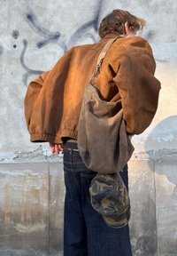 Brun bomberjakke i ruskind med oversize ærmer, mørk denim jeans og en grå skuldertaske i ruskind, der holder en camouflage-kasket.