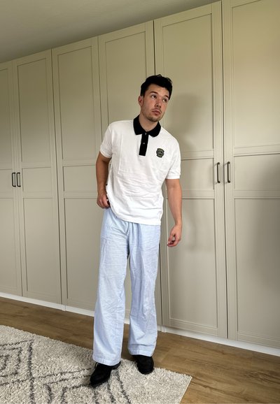 Camisa polo blanca con cuello negro y emblema, combinada con pantalones a rayas azul claro y zapatos negros. De pie frente a un fondo neutro.