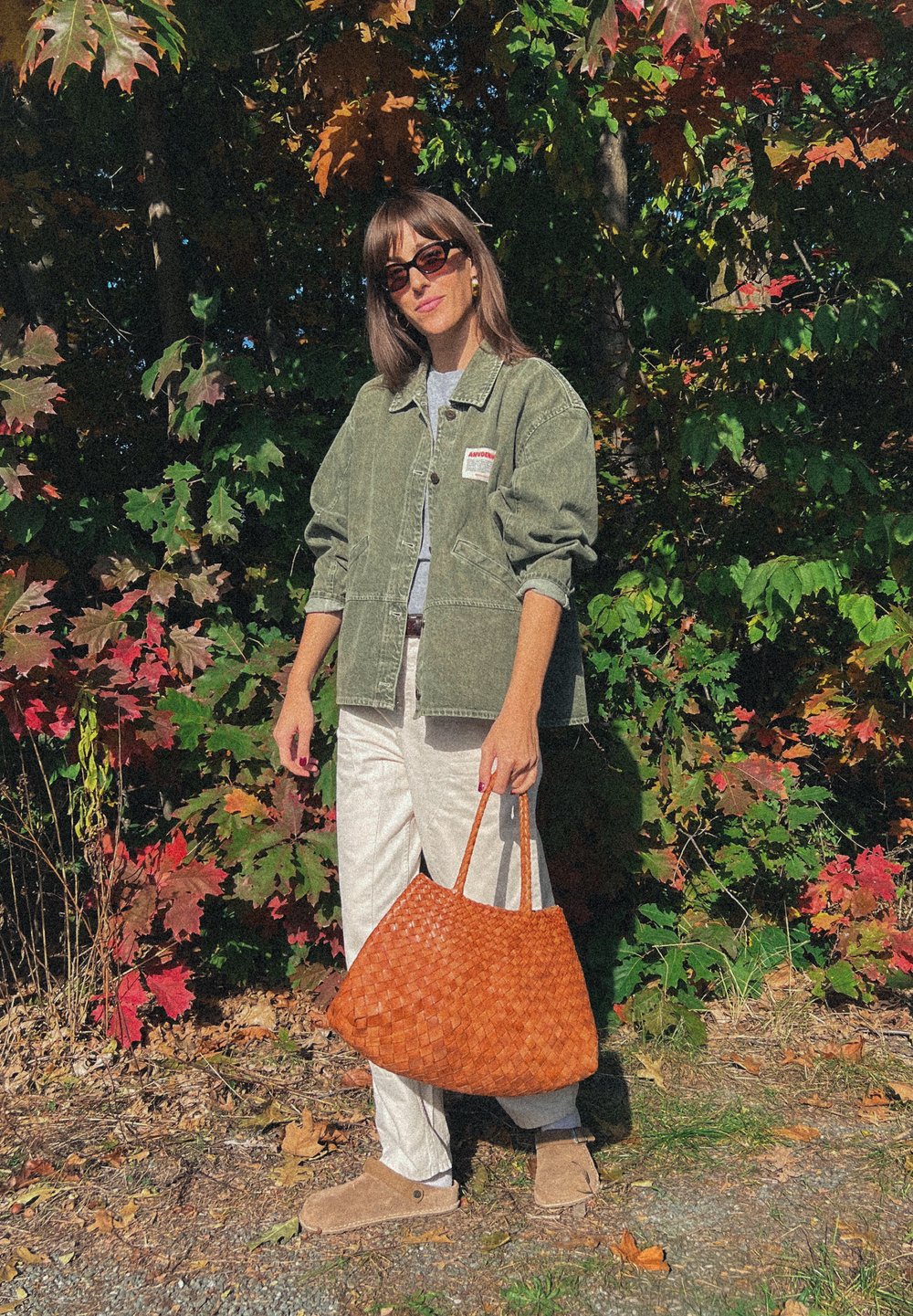 Giacca in corda verde oliva oversize, t-shirt grigia, pantaloni bianchi, borsa intrecciata marrone e stivali alla caviglia beige contro un sfondo di colorate foglie autunnali.