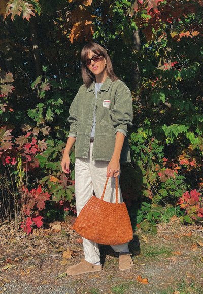 Chaqueta de corduroy verde oliva de gran tamaño, camiseta gris, pantalones blancos, bolso de mano tejido marrón y botines beige frente a un colorido follaje otoñal.