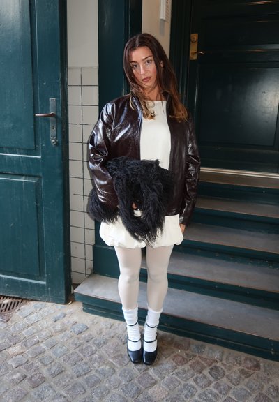 Chaqueta bomber de cuero marrón, vestido blanco, bolso de piel sintética negro, medias claras y zapatos negros con calcetines blancos. Fondo con puerta azul-verde.