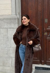 Manteau en fausse fourrure marron avec un large col, associé à un haut marron et un jean bleu clair. Tenant un petit sac à main marron foncé. Porte en bois en arrière-plan.