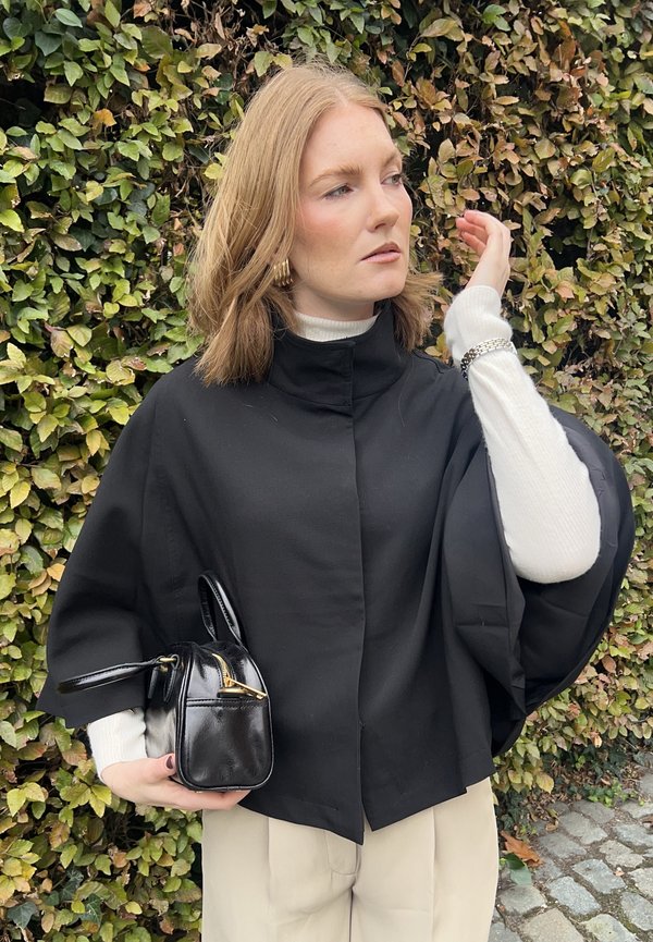 Cazadora ligera negra estilo capa con un cuello alto, combinada con un cuello de tortuga blanco estriado. Sosteniendo un pequeño bolso negro con detalles dorados.