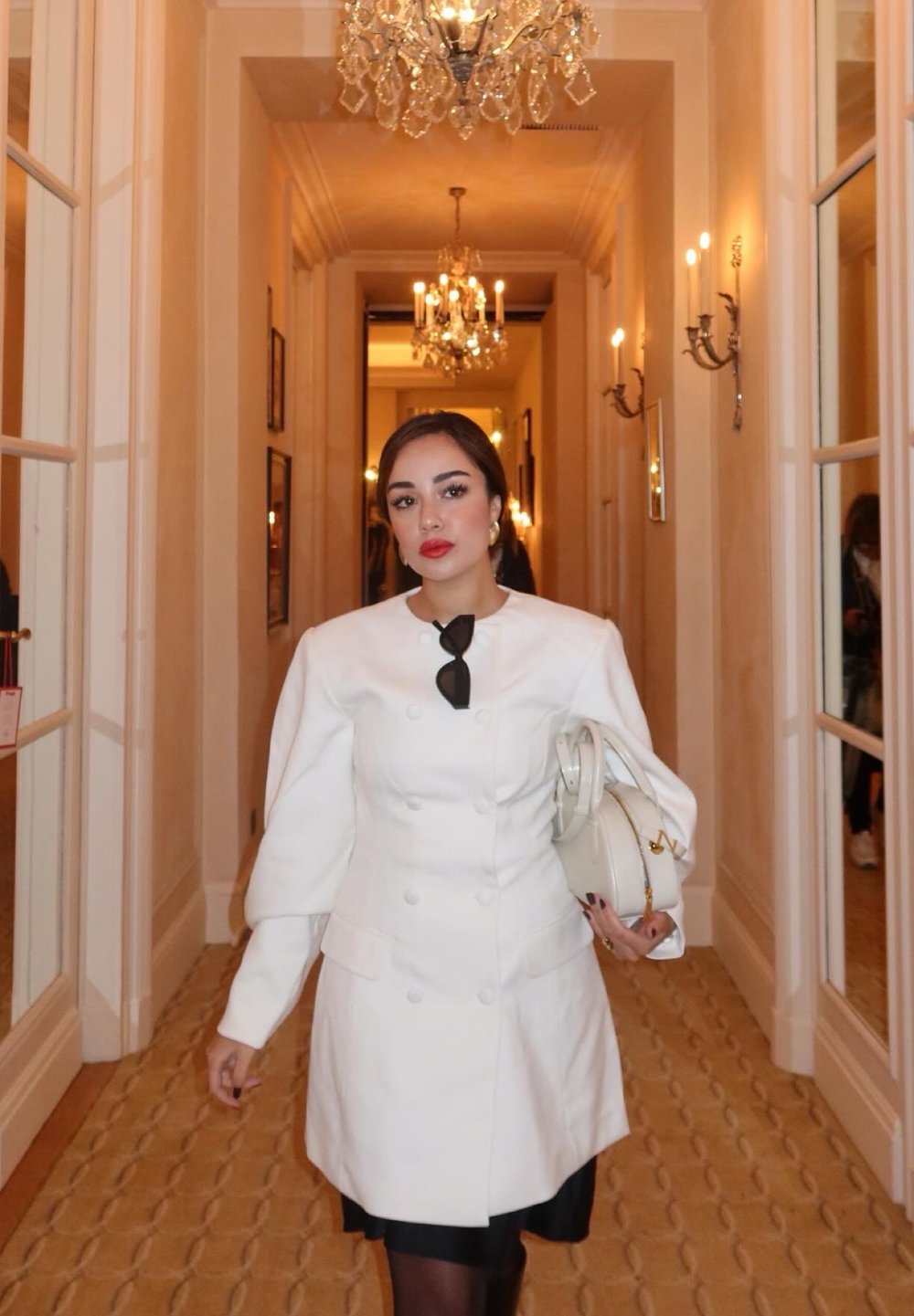 Cappotto bianco doppiopetto con maniche a sbuffo, abbinato a un vestito nero. Porta una borsa bianca. Hall elegante con lampadari sullo sfondo.