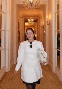 Cappotto bianco doppiopetto con maniche a sbuffo, abbinato a un vestito nero. Porta una borsa bianca. Hall elegante con lampadari sullo sfondo.