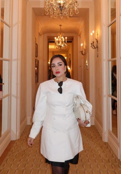 Abrigo blanco de doble botonadura con mangas abullonadas, combinado con un vestido negro. Lleva un bolso blanco. Pasillo elegante con candelabros al fondo.