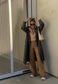 Overmaatse donkergroene trenchcoat, beige geribde trui, tan wijde broek en witte sneakers met paarse accenten. Zonnebril en een witte handtas.