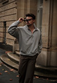 Sivá mikina na zips s uvoľneným strihom, nosená cez biele tielko, doplnená o hnedé nohavice. Okrúhly golier a ležérny štýl.