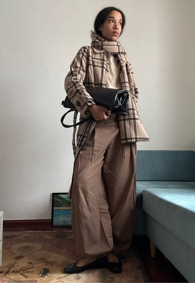 Joven con abrigo beige a cuadros y bufanda, pantalones anchos marrones, zapatos planos negros, sosteniendo un gran bolso de mano negro, de pie en el interior cerca de un sofá.