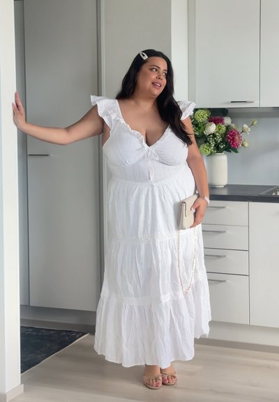 Mujer con un vestido blanco de capas, sujetando un clutch beige, de pie en una cocina moderna cerca de un jarrón con flores coloridas.