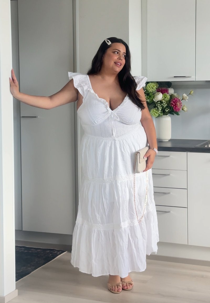 Femme en robe blanche à étages tenant une pochette beige, debout dans une cuisine moderne près d’un vase avec des fleurs colorées.