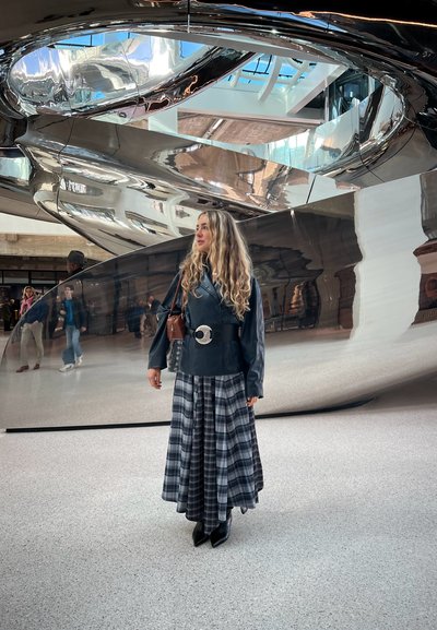 Mujer con cabello largo y rizado, vestida con chaqueta de cuero negra y falda de cuadros, de pie frente a una gran escultura metálica reflectante en interiores.