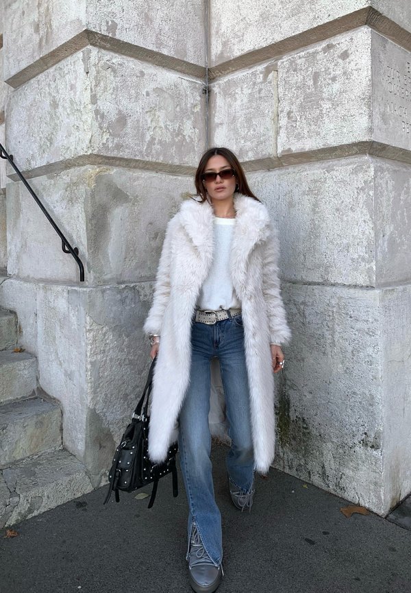 Weißer Kunstpelz-Mantel mit Schalkragen, leichter Pullover, ausgestellte blaue Jeans, silberne Stiefel und eine schwarze Nieten-Handtasche vor einer Steinmauer.