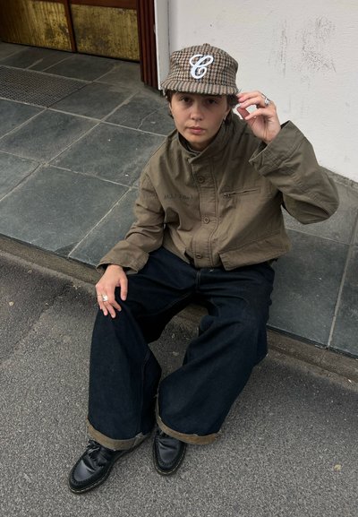 Persona con gorra estampada, chaqueta oliva, vaqueros anchos y oscuros y zapatos negros sentada en una acera cerca de la pared de un edificio.