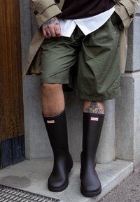Bottes de pluie en caoutchouc marron avec une texture lisse et des semelles prononcées, arborant une marque rouge et blanche sur le dessus. Associées à un short vert olive.