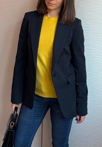 Γυναίκα που φοράει μαύρο σακάκι πάνω από κίτρινο πουλόβερ, μπλε τζιν, κρατώντας μια μαύρη τσάντα χειρός μπροστά σε έναν υφασμάτινο μπεζ και λευκό τοίχο.