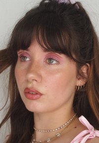 Portrett av en person med langt, bølgete brunt hår, iført gulløredobber og lagdelte halskjeder. De har rosa øyeskygge og en hjerteformet næse piercing.
