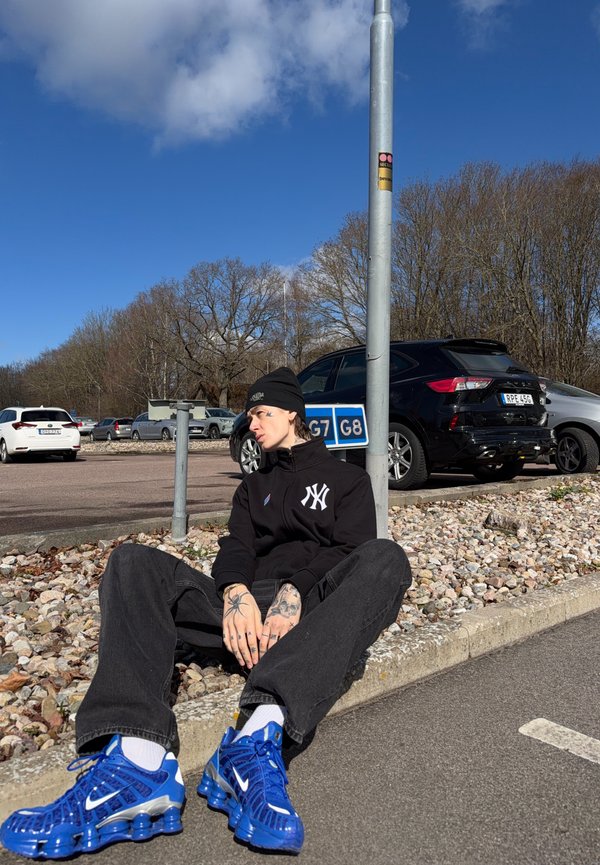 Young person with tattoos wearing black hat, black jacket, and bright blue Nike sneakers sitting on curb in parking lot under clear sky.