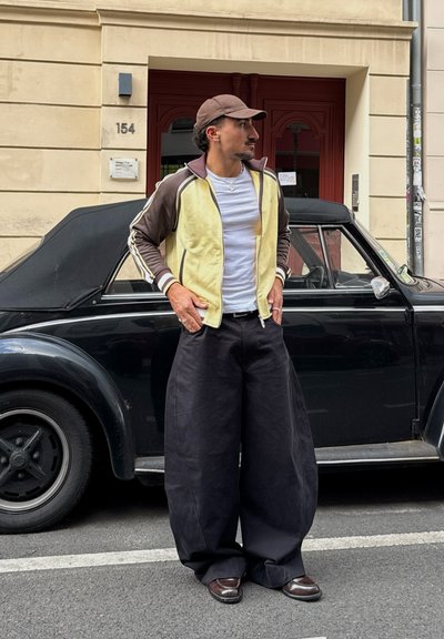 Chaqueta de chándal amarilla y marrón sobre una camiseta blanca, combinada con pantalones anchos negros y zapatos marrones, con un fondo de un coche convertible negro.