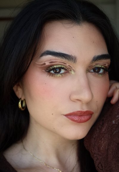 Mujer con cabello oscuro que lleva pendientes de aro dorados y sombras de ojos verde oliva con patrones abstractos marrones, posando con la mano cerca del rostro.