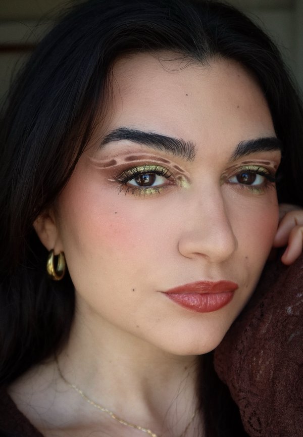 Femme aux cheveux sombres portant des boucles d'oreilles créoles dorées et un fard à paupières vert olive avec des motifs abstraits marron, posant la main près du visage.