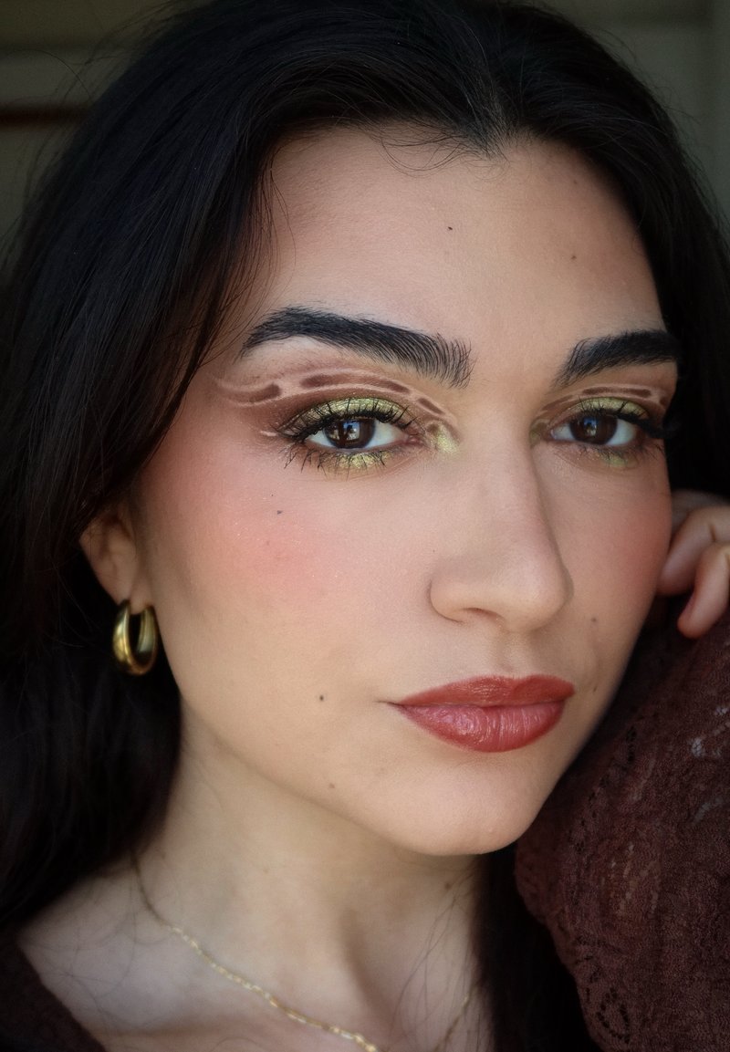 Femme aux cheveux foncés portant des boucles d'oreilles en or en forme d'anneau et un fard à paupières vert olive avec des motifs abstraits bruns, posant avec la main près du visage.