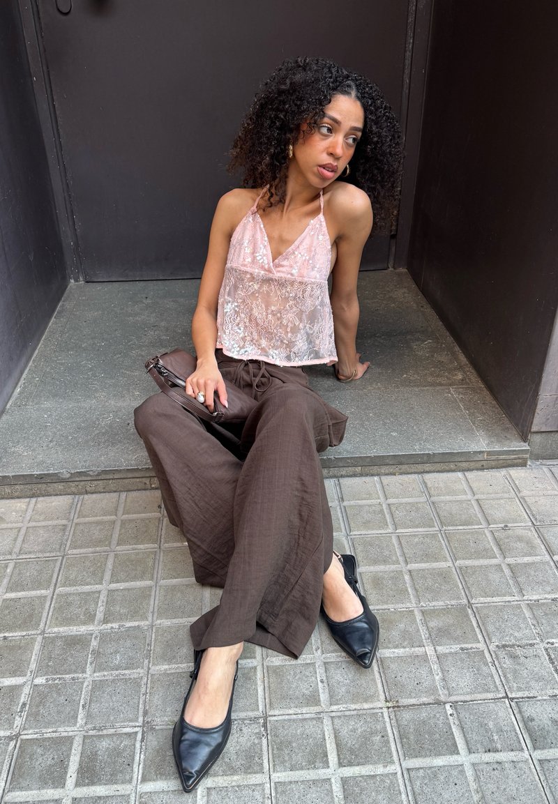 Femme aux cheveux bouclés assise sur une marche, portant un haut rose en dentelle à licou, un pantalon large marron et des ballerines noires, regardant sur le côté.