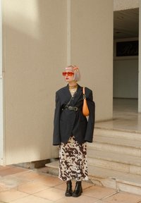 Overdimensioneret sort blazer, cremefarvet kjole med brune pletter, orange håndtaske, klare røde solbriller, sorte klodsede støvler, lagdelte guldkæder.