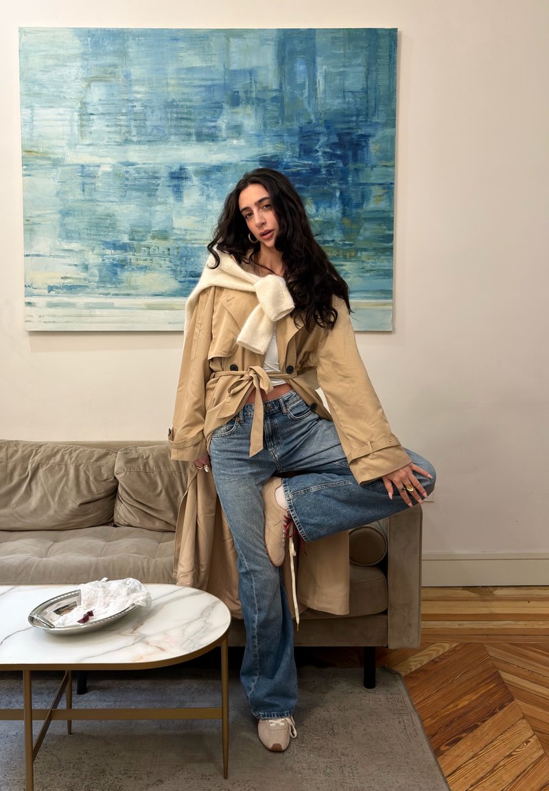 Femme aux longs cheveux foncés, portant un trench-coat beige et un jean bleu, posant avec une jambe sur un canapé beige sous une grande peinture abstraite bleue.