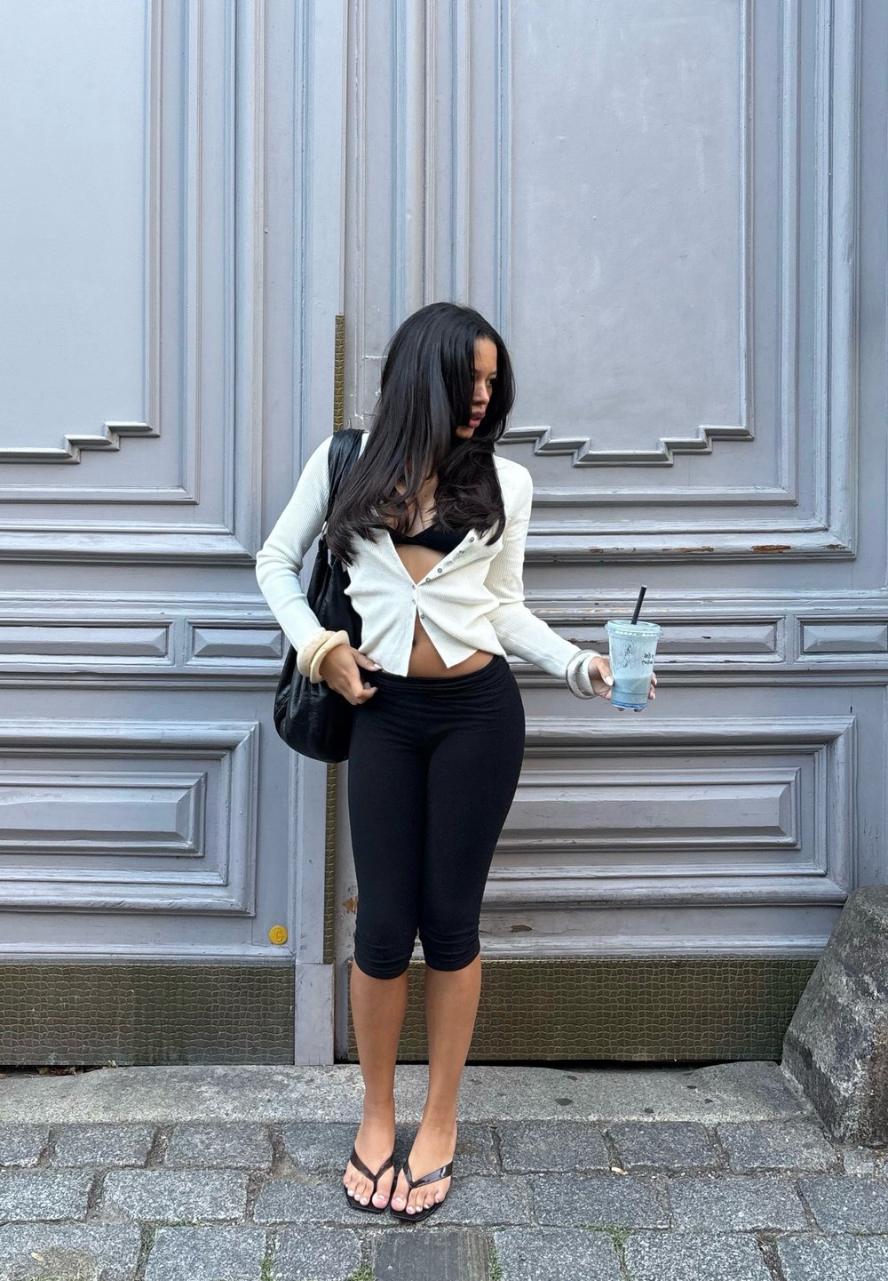 Biely rebrovaný kardigán, čierny crop top, čierne legíny a čierne žabky. Drží priehľadný nápoj. Pozadie s šedými panelmi.