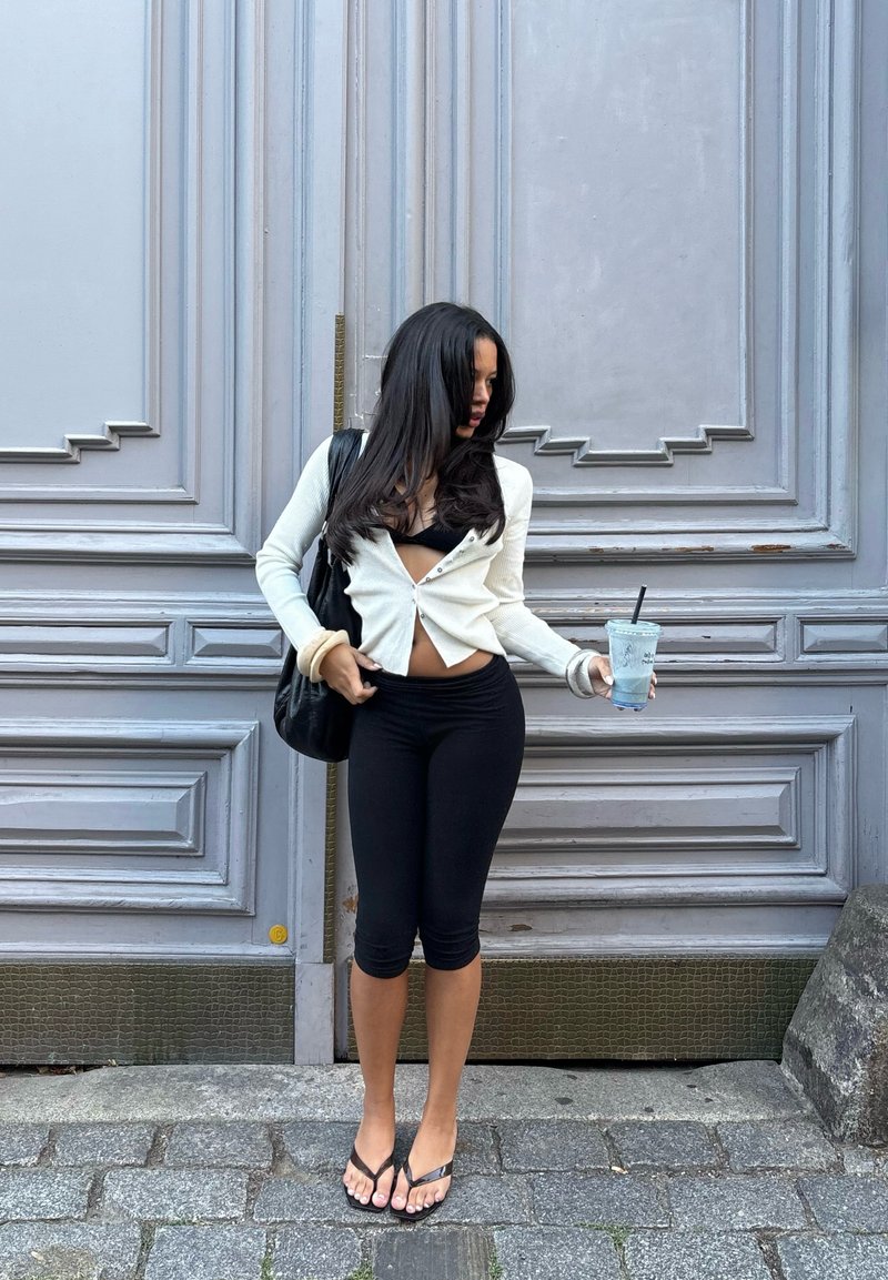 Weisser gerippter Cardigan, schwarzes Crop-Top, schwarze Leggings und schwarze Flip-Flops. Hält ein klares Getränk in der Hand. Im Hintergrund sind graue Paneele zu sehen.