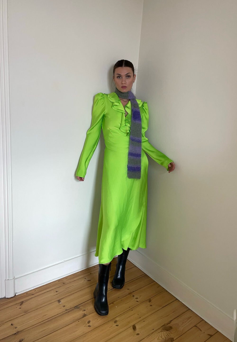 Vestido verde brilhante com detalhes em babados, mangas longas e uma silhueta fluida, combinado com um lenço roxo riscado e botas pretas até ao tornozelo.