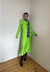 Vestido verde brilhante com detalhes em babados, mangas longas e uma silhueta fluida, combinado com um lenço roxo riscado e botas pretas até ao tornozelo.
