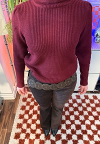 Personne portant un pull en maille bordeaux, un pantalon foncé et une ceinture avec de grandes plaques métalliques circulaires, debout sur un tapis à carreaux rouges et blancs.