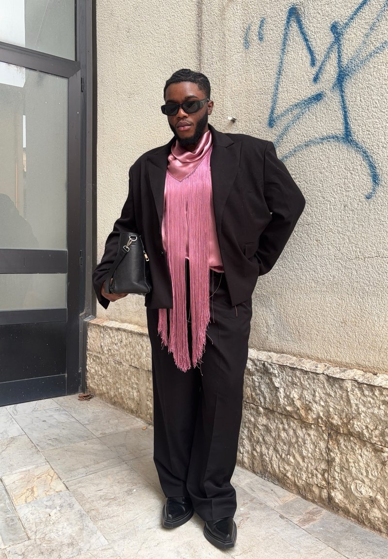 Black suit jacket over a pink satin shirt featuring fringe detail. Accompanied by black trousers and a bag. Worn with black sunglasses and shoes. Textured background.