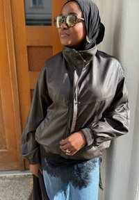 Tamno smeđa kožna jakna s visokim ovratnikom, zatvaranjem na zatvarač i rebrastim manžetama. Uparena s svijetloplavim trapericama s čipkastim rubovima.